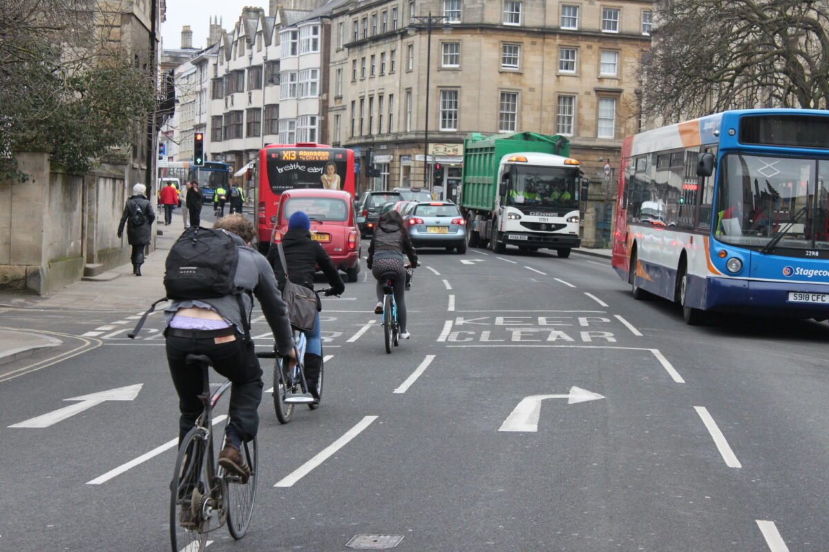 Three people cycling in the cycle lane filter on Magdalen Bridge, Oxford, in the direction of the High Street to turn right onto Longwall Street. The road is shared by busses, cars and a lorry.