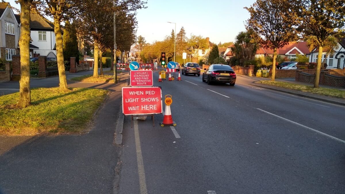 Roadwork signs block the painted cycle lane at a set of temporary traffic lights on a wide road. A smaller red sign above a larger red sign advising the traffic to stop when the light shows red says: 'cyclists's dismount and find an alternative route'.