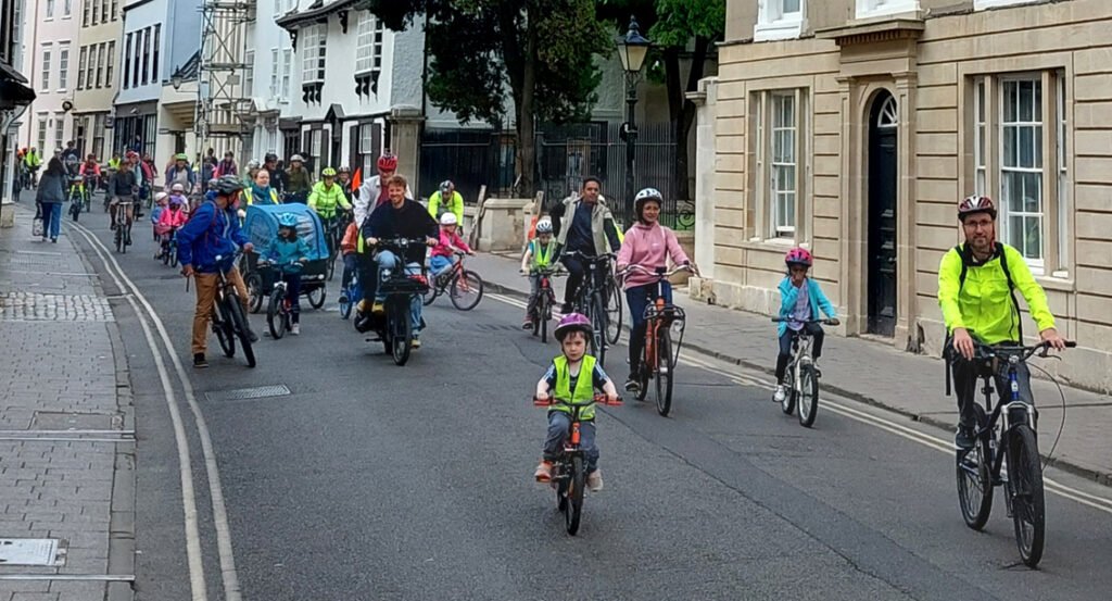 Large diverse group of cyclists, adults and children, riding down Holywell Street, Oxford