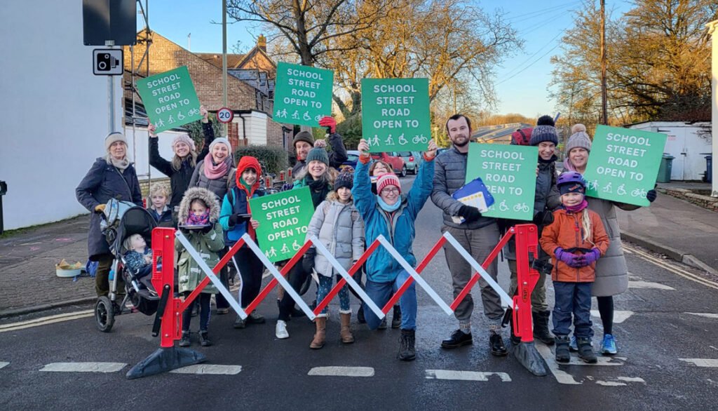 Group of adults and children holding placards standing at entrance to side road blocked by a temporary barrier