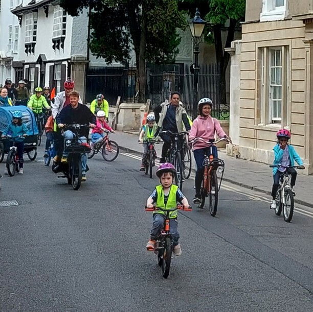 Large diverse group of cyclists, adults and children, riding down Holywell Street, Oxford