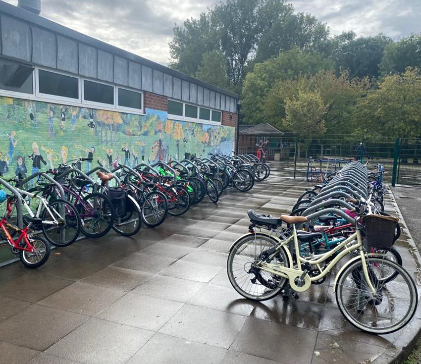 Crowded cycle racks on wet pavement next to school wall painted with mural.