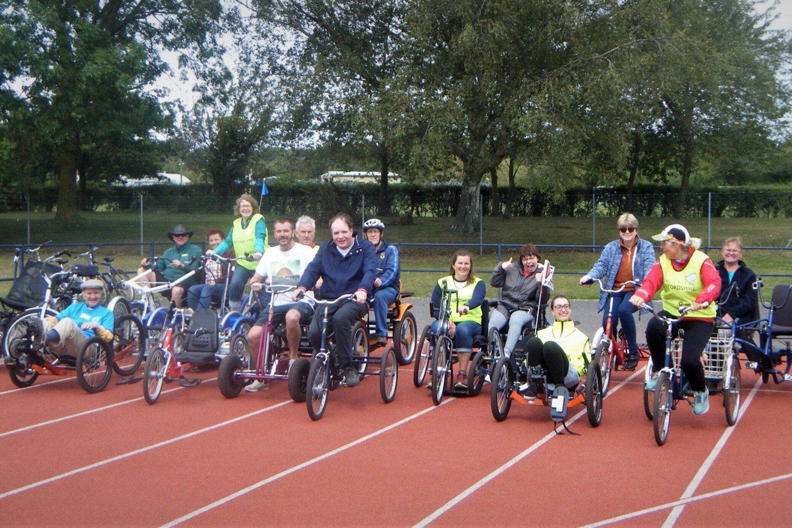 Group of cyclists of all abilities on a range of different types of bike, including bicycles, tricycles, low-slung four-wheelers, hand powered. Lined up on an athletics track