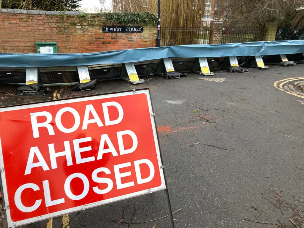 Sign saying 'Road ahead closed' in foreground. Behind it flood barriers 