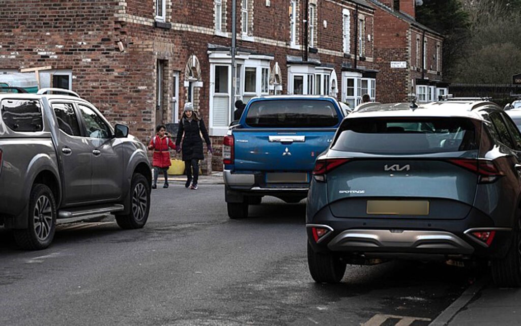 A residential street crammed with 4x4 vehicles, parked on the pavement and clogging the road. A woman and child walking past the cars