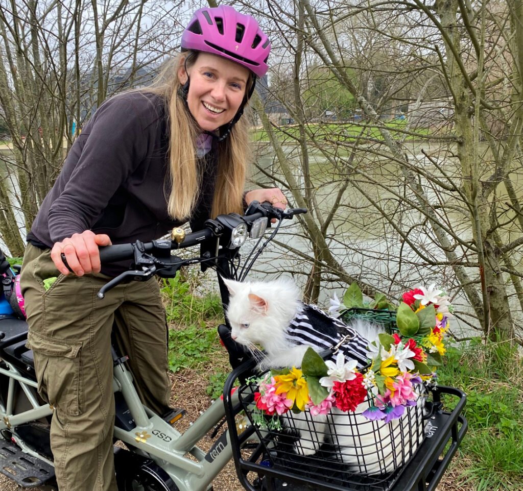White woman seated on stationary e-bike leans forward to look into camera. On the front of her bike is a black wire basket decorated with artificial flowers and containing a fluffy white cat wearing a harness.