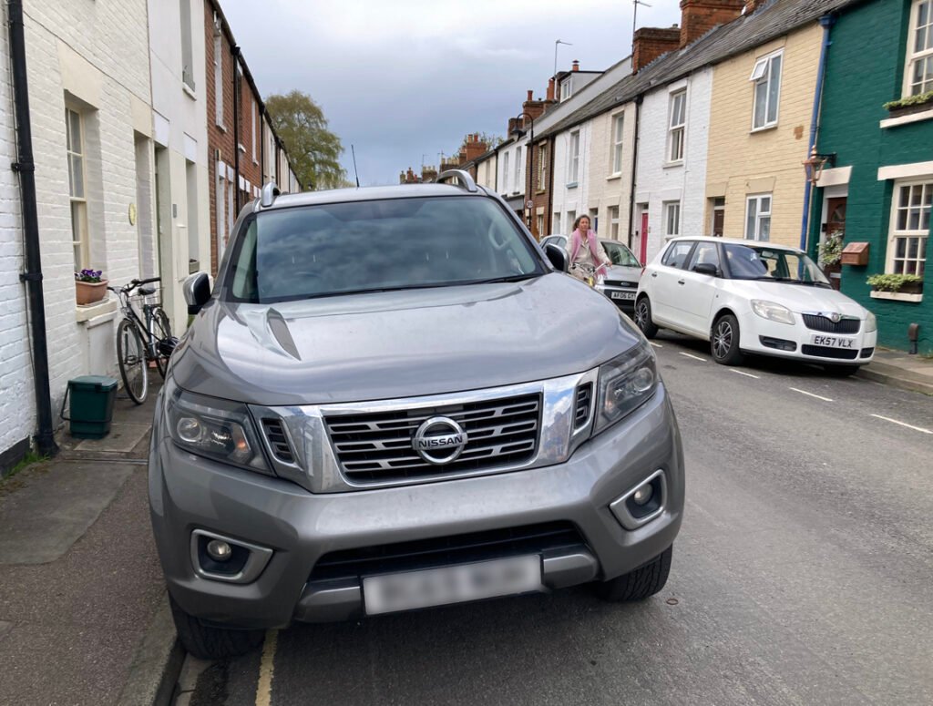 Grey SUV parked in narrow residential street of terraced housing. A cyclist is coming up behind the car but partly obscured by the vehicle