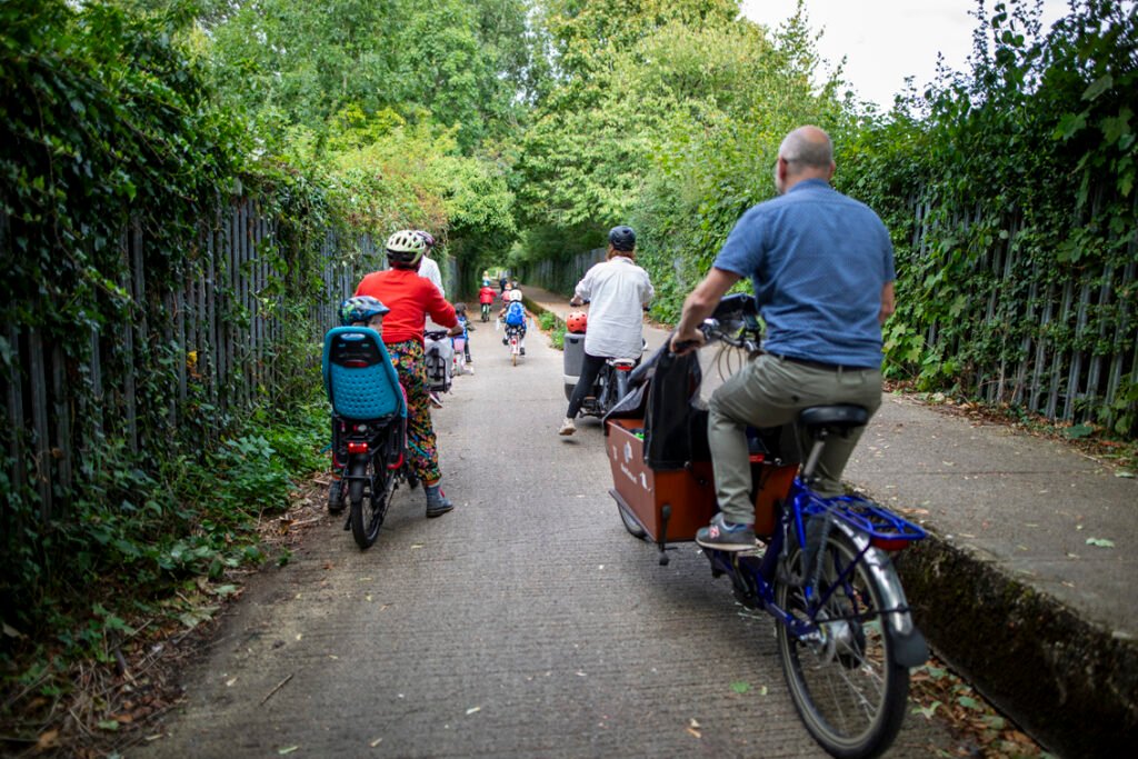 Narrow path bordered by greenery and fences with mixed-age cyclists