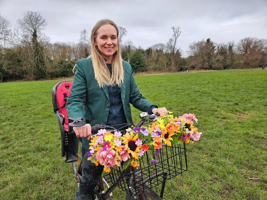 White woman with long blonde hair posing in a field with bike that has child seat and front basket decorated with plastic flowers