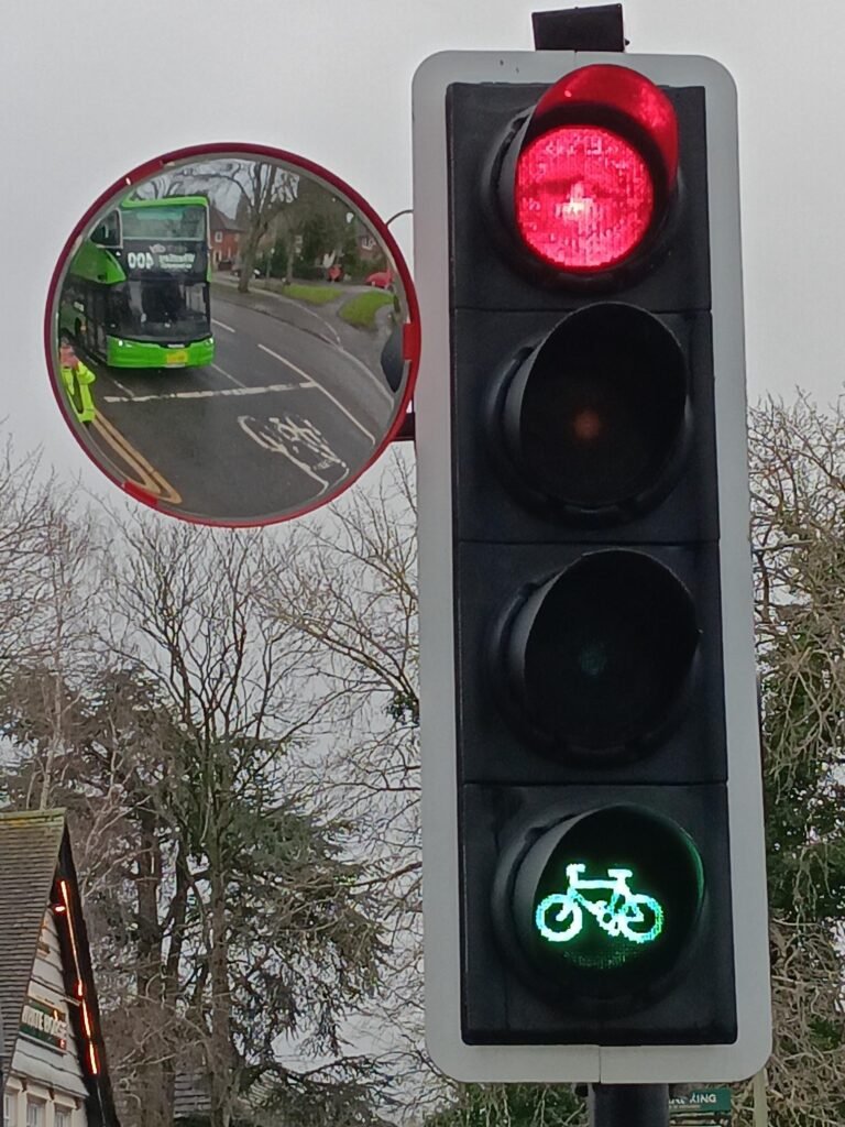 Traffic light on red, with green bike filter light. Convex mirror attached to light reflects bus stopped at the light and bicycle in bike lane